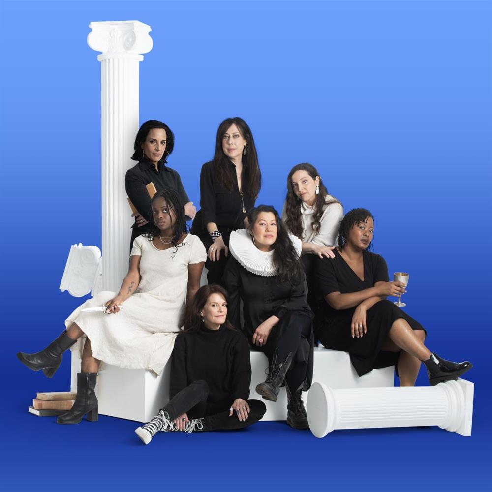 Seven women of various ages and ethnicities sit on white blocks against a blue backdrop, staring at the viewer.